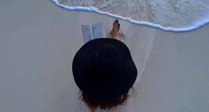 a lighter skinned person sitting on the beach near water and reading with a big hat on.jpg.optimal