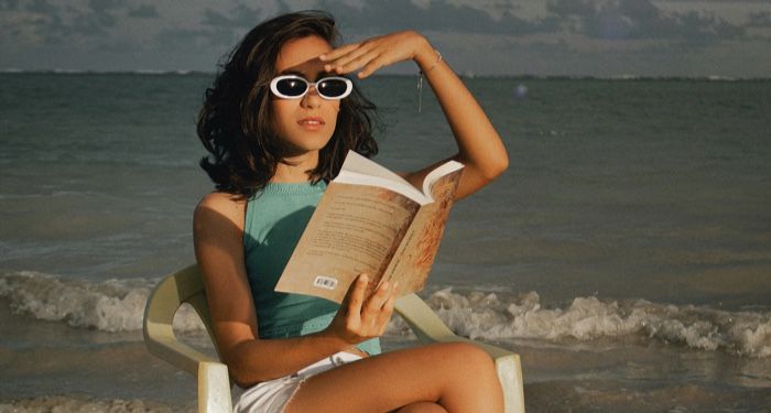 a lightly tanned skin woman reads a book while sitting in s a white chair on the beach.jpg.optimal