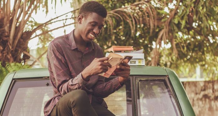 a dark brown skinned Black man smiling and reading a book while sitting on a jeep with palm trees in the back.jpg.optimal