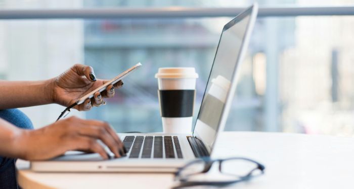 woman holding phone with hands on laptop keyboard in an office with a to go coffee on eyeglasses on desk feature.jpg.optimal